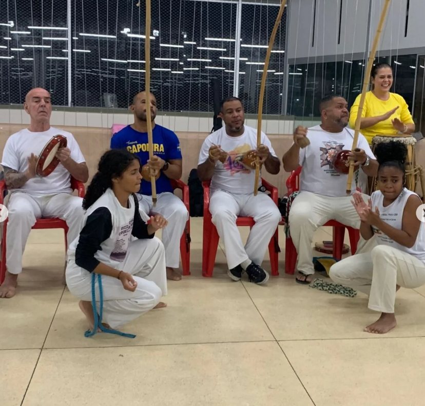 Alunos tocando berimbau e pandeiro sentados em roda